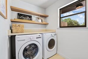 A laundry nook with a full-size white front-loading washer and dryer beneath a beige countertop keeps the long ranch weekend running. Two wood floating shelves hold a basket, a black laundry sign, and decor above a window onto the wood fence.
