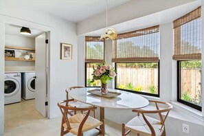 A bright breakfast nook with an oval white marble-look table and two wood wishbone chairs sits beside a bay of black-framed windows under bamboo shades. An open doorway leads straight into the laundry room with white washer and dryer side by side.
