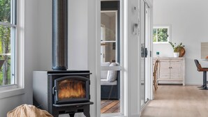 A modern living room with a wood-burning stove, a large window, and a view of the outdoors. - "Sunset Valley Views" – Close to Beach (Avoca Beach)