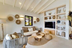 A living room with a sofa, chairs, a coffee table, and a TV. There are wall decorations and a shelf with various items.