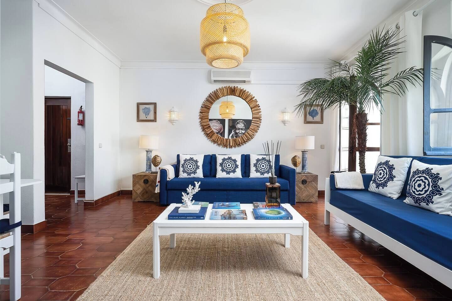 A modern living room with a blue sofa, a coffee table, and a large mirror. There are two side tables with lamps, a potted plant, and a door leading to another room.