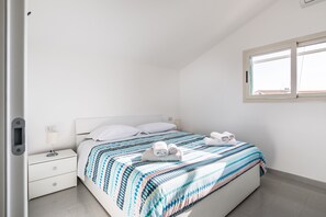 A neatly made bed with a patterned bedspread, a bedside table with a lamp, and a window with blinds. - Artemide Vista Mare (Ragusa)