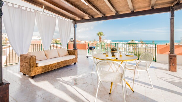 Un área de estar al aire libre con techo, con un sofá de mimbre y sillas blancas, una mesa redonda con cubierta amarilla y vista al mar.