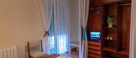 A bedroom with a bed, a chair, a desk with a computer, a wooden wardrobe, and a window with curtains.