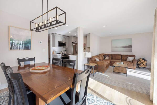 A modern living room with a wooden dining table, black chairs, a brown leather sofa, and a coffee table. There is a kitchen area with white cabinets and a microwave.