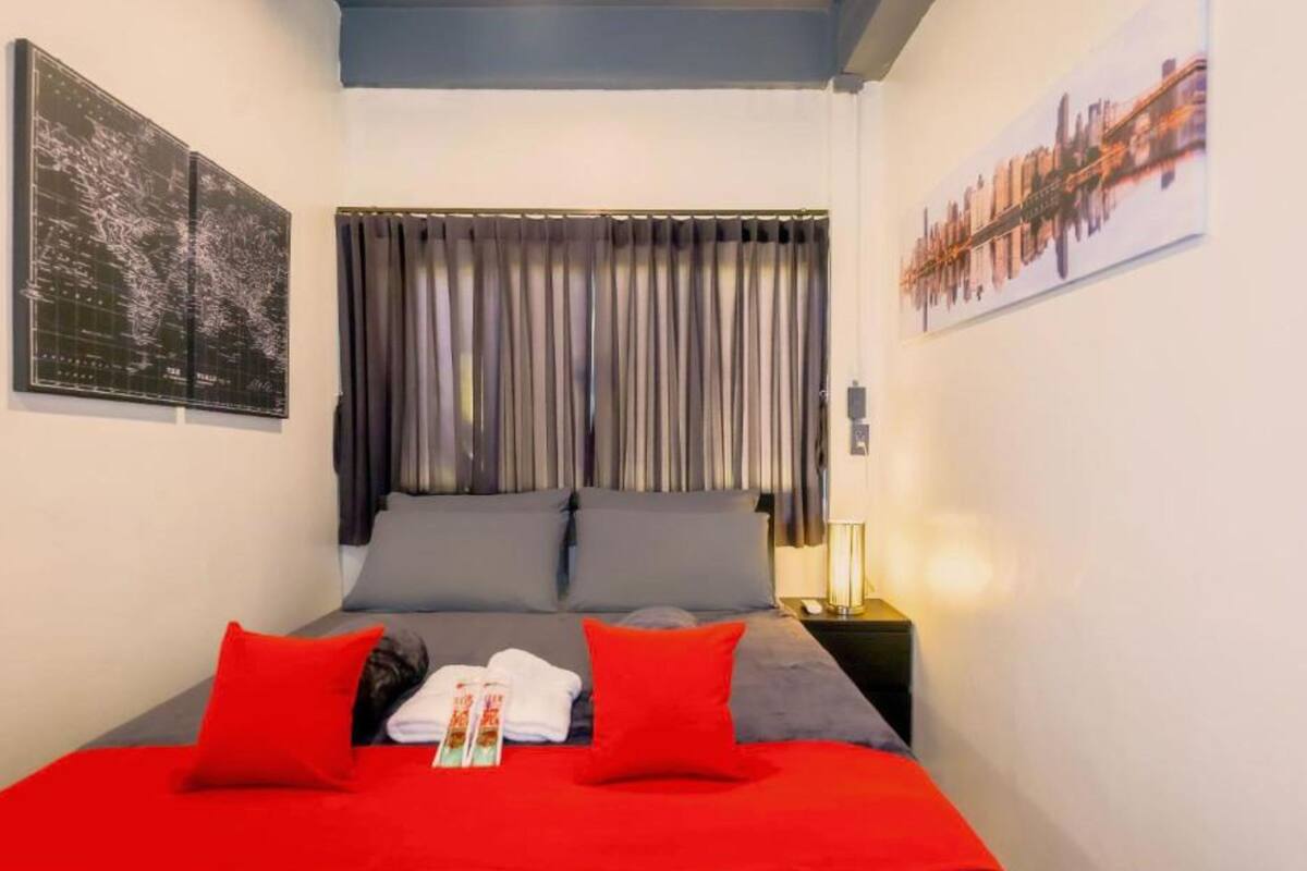 Una cama bien hecha con ropa de cama roja y gris, un mapa en la pared y un cuadro de un paisaje urbano.