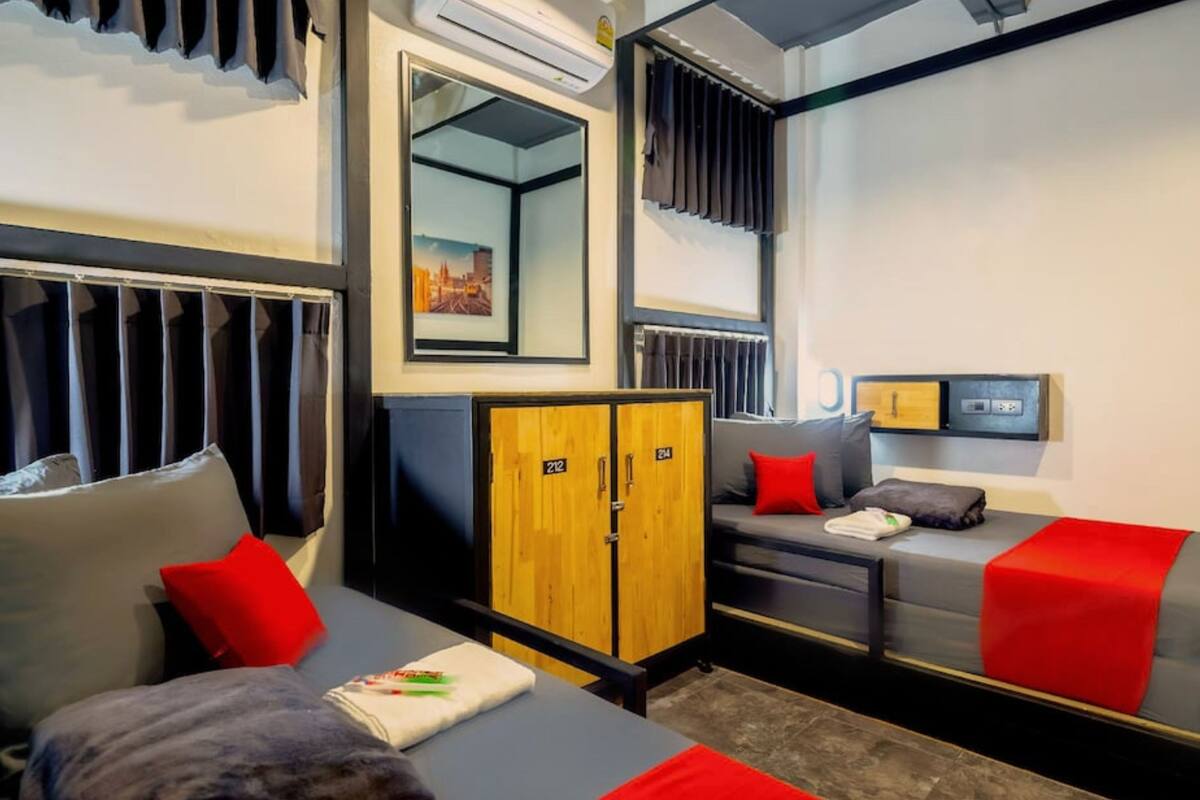 Una habitación de hotel moderna con sofá gris, cojines rojos, armario de madera y un espejo que refleja un paisaje urbano.