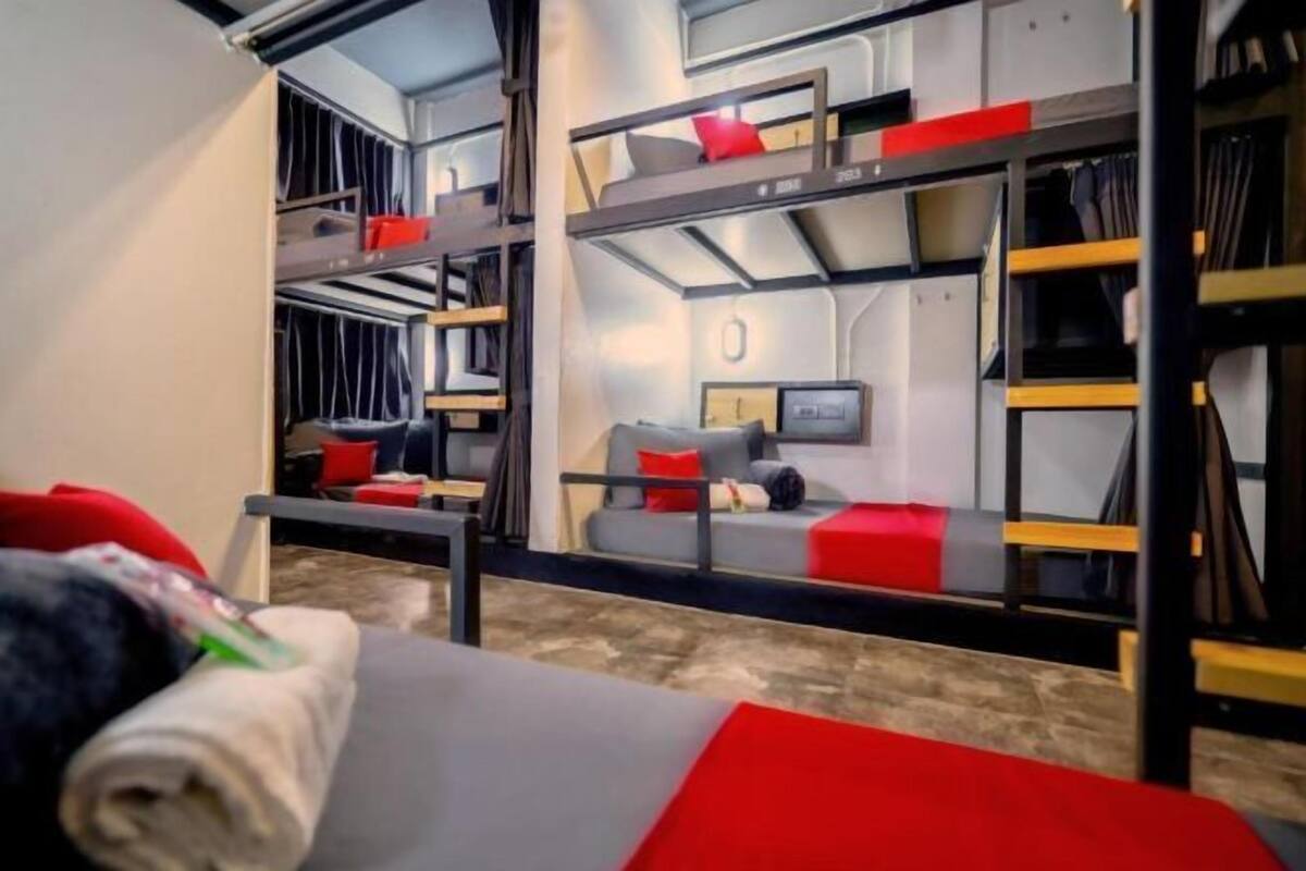 Habitación con literas, un sofá gris, cojines rojos y una mesita pequeña.