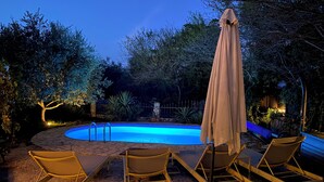 Zona de piscina con sillones de descanso y una sombrilla, rodeada de árboles y un sendero de piedra.