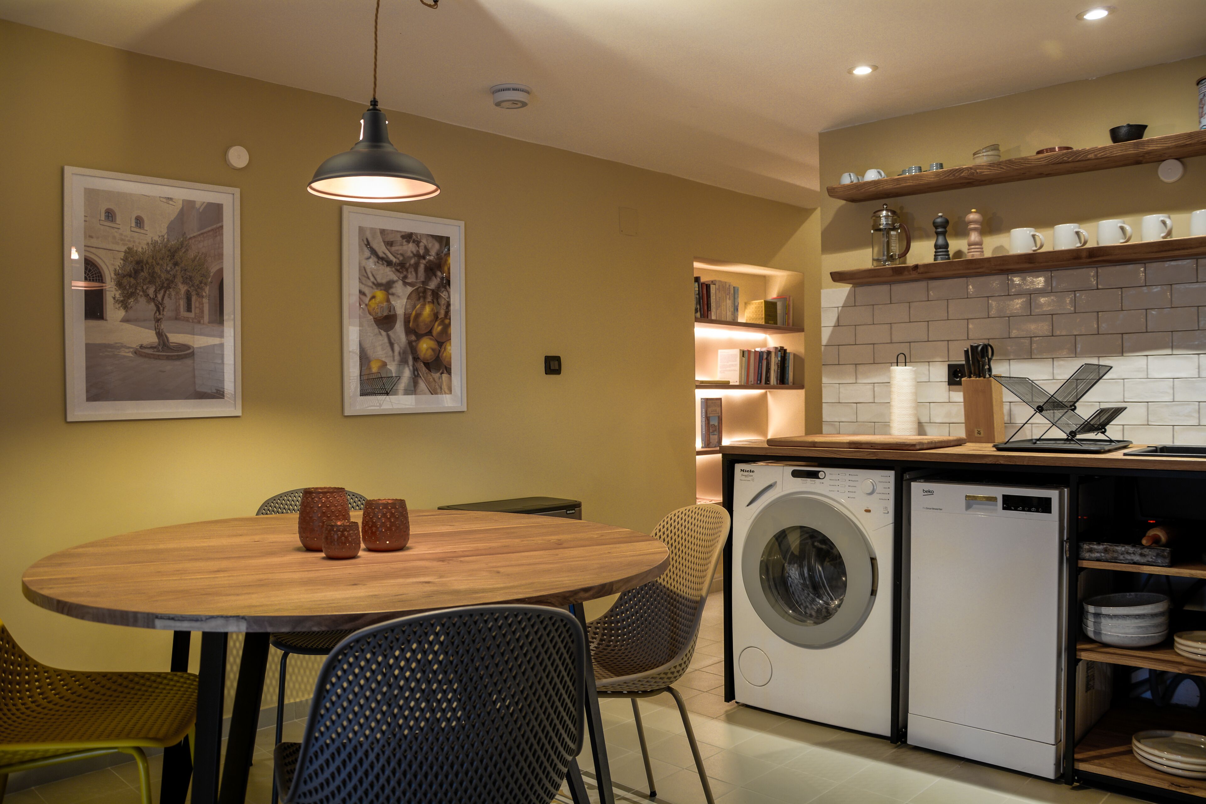 A compact kitchen with a wooden dining table, chairs, a washing machine, and a shelf with dishes.