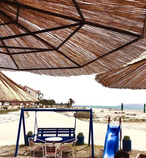 A beachside seating area with a swing set, a slide, and umbrellas providing shade.