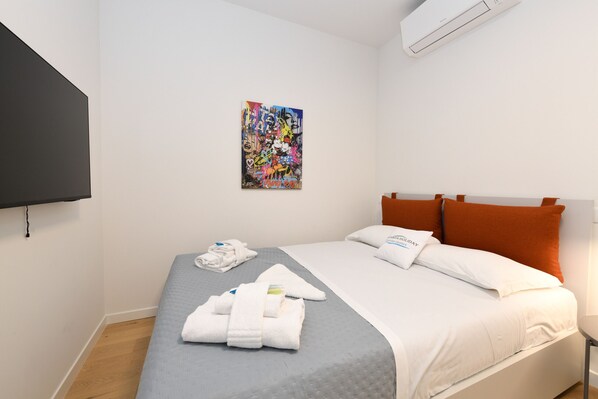 Une chambre d’hôtel avec un lit, un téléviseur à écran plat fixé au mur, une œuvre d’art colorée au-dessus du lit et une paire d’oreillers orange.