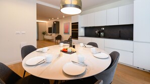 Een moderne eethoek met een ronde tafel voor vier personen, witte borden, bestek en een waterfles. Daarboven hangt een hanglamp en de kamer heeft witte kasten en een zwart aanrecht.