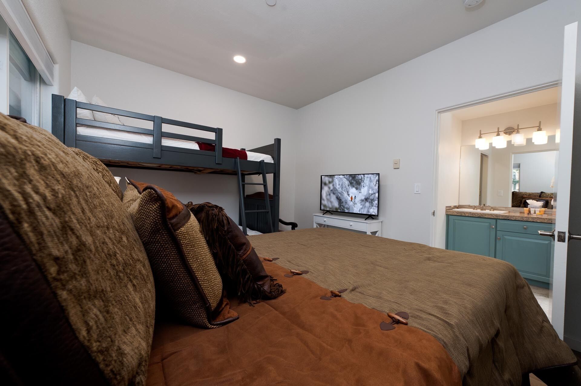A room with bunk beds, a television, a bathroom with a mirror and sink, and a bed with a brown blanket and pillows.