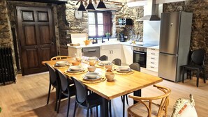 Un comedor con una mesa de madera dispuesta para cuatro personas, rodeada de sillas. Se ve una cocina con refrigerador, horno y pileta al fondo.