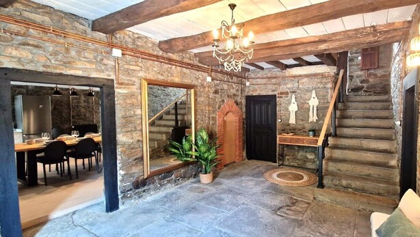 Un interior rústico con paredes de piedra vista, una escalera de madera, una zona de comedor con mesa y sillas, y una araña.