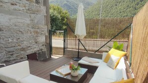 Una terraza con pared de piedra, asientos blancos acolchados, una mesa con un jarrón y vasos, y vistas a colinas verdes.