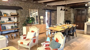 Un comedor rústico con una mesa de madera dispuesta para cuatro, rodeada de sillas tapizadas con cojines estampados. Se aprecia una pared de piedra y vigas de madera al fondo.