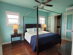 Un lit à baldaquin avec une structure en bois et une couvre-lit bleu foncé, flanqué de deux tables de chevet avec des lampes, dans une pièce aux murs de couleur turquoise et dotée d’un ventilateur de plafond.