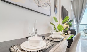 Une table Ă manger dressĂ©e avec de lâassiette, des bols et des verres blancs, une plante et une vue sur la ville Ă travers de grandes fenĂȘtres.