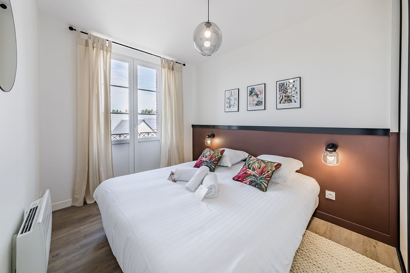 Ein ordentlich bezogenes Bett mit weißen Leinen und dekorativen Kissen, ein Nachttisch mit Lampe und Blick ins Freie durch ein großes Fenster mit Vorhängen.