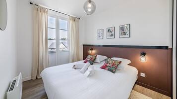 Ein ordentlich bezogenes Bett mit weißen Leinen und dekorativen Kissen, ein Nachttisch mit Lampe und Blick ins Freie durch ein großes Fenster mit Vorhängen.