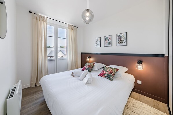 A neatly made bed with white linens and decorative pillows, a bedside table with a lamp, and a view of the outdoors through a large window with curtains.