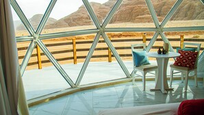 A room with a large glass window offering a view of a desert landscape. Inside, there is a white round table with two chairs, a small rug, and a bed with a red pillow. - Desert Season Camp (Wadi Rum)