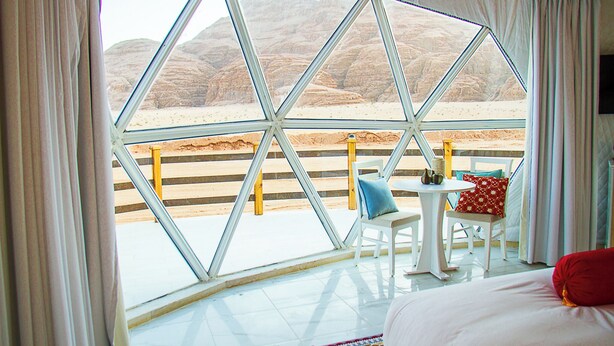 A room with a large window offering a view of a desert landscape. Inside, there is a bed with a red pillow, a small table with a blue chair, and a white chair. - Desert Season Camp (Wadi Rum)