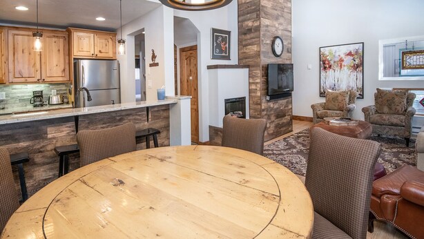 A modern kitchen with wooden cabinets, a stainless steel refrigerator, and a central island. There is a dining area with a round wooden table and two chairs. The living room features a fireplace, a flat-screen TV, and a large window with artwork.