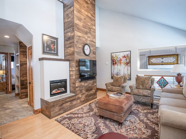 Una sala de estar con chimenea, televisor montado en la pared, un cuadro grande y una zona de estar acogedora con alfombra.