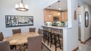 A dining area with a wooden table and chairs, a kitchen with wooden cabinets, and a hanging light fixture.