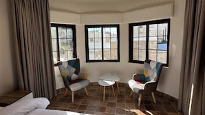 A hotel room with a bed, two armchairs, a small table, and three windows.