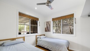 Une chambre avec deux lits simples, un plancher en bois, des murs blancs et de grandes fenêtres équipées de stores.