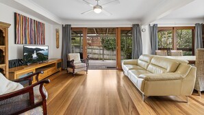 Un salon spacieux doté d’un plancher en bois, d’un canapé beige, d’un meuble de rangement en bois abritant un téléviseur, et offrant une vue sur un jardin grâce à des portes vitrées.