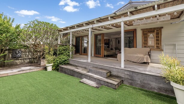 Un patio couvert avec une terrasse en bois, un petit jardin agrémenté de gazon synthétique et une vue sur l’intérieur de la maison à travers des portes vitrées.