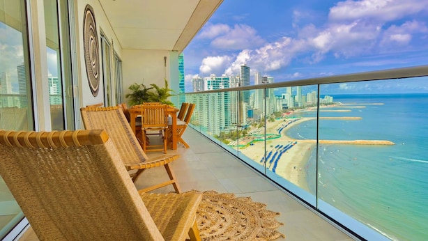A balcony with wicker furniture, a view of a beach, and high-rise buildings in the background. - Apartamento Lujoso H2 - Bocagrande (Cartagena)