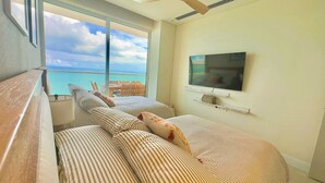 A room with a large window overlooking the sea, a bed with striped bedding, a television mounted on the wall, and a small shelf with decorative items. - Apartamento Lujoso H2 - Bocagrande (Cartagena)