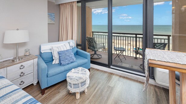 A hotel room with a blue sofa, a small round ottoman, a wooden nightstand, a lamp, and a view of the beach through large glass doors. - Oceanfront King Suite - Fabulous Views - King Bed - Indoor and Outdoor pools (Myrtle Beach)