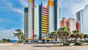 A multi-story hotel with a modern design, featuring a mix of colors on the facade and a parking area with palm trees. - Oceanfront King Suite - Fabulous Views - King Bed - Indoor and Outdoor pools (Myrtle Beach)