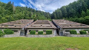 A row of three-story buildings with stone facades and flat roofs, surrounded by a well-maintained lawn and dense forest. - Happy Guest Apartments Mountain Panorama Borno (BORNO)