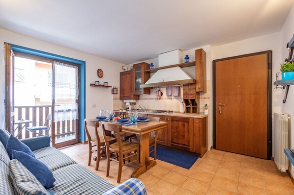 Cocina pequeña con armarios de madera, una mesa de comedor con individuales azules y una puerta corrediza con marco azul que da a un balcón.