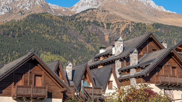 A row of traditional wooden chalets with steep roofs, set against a backdrop of a mountainous landscape.