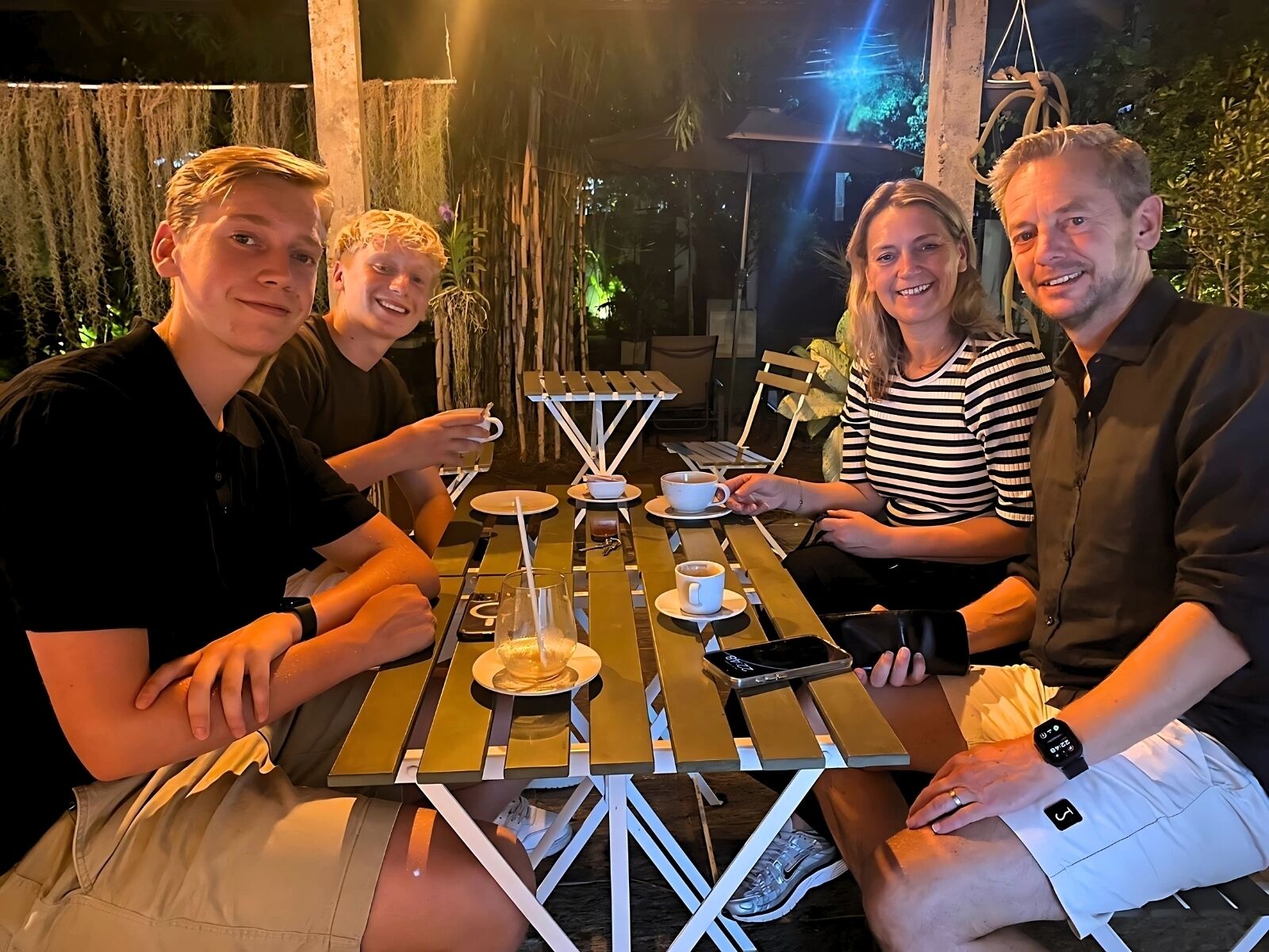 Un grupo de personas sentadas en una mesa al aire libre con una taza de café, un teléfono inteligente y un reloj sobre la mesa.