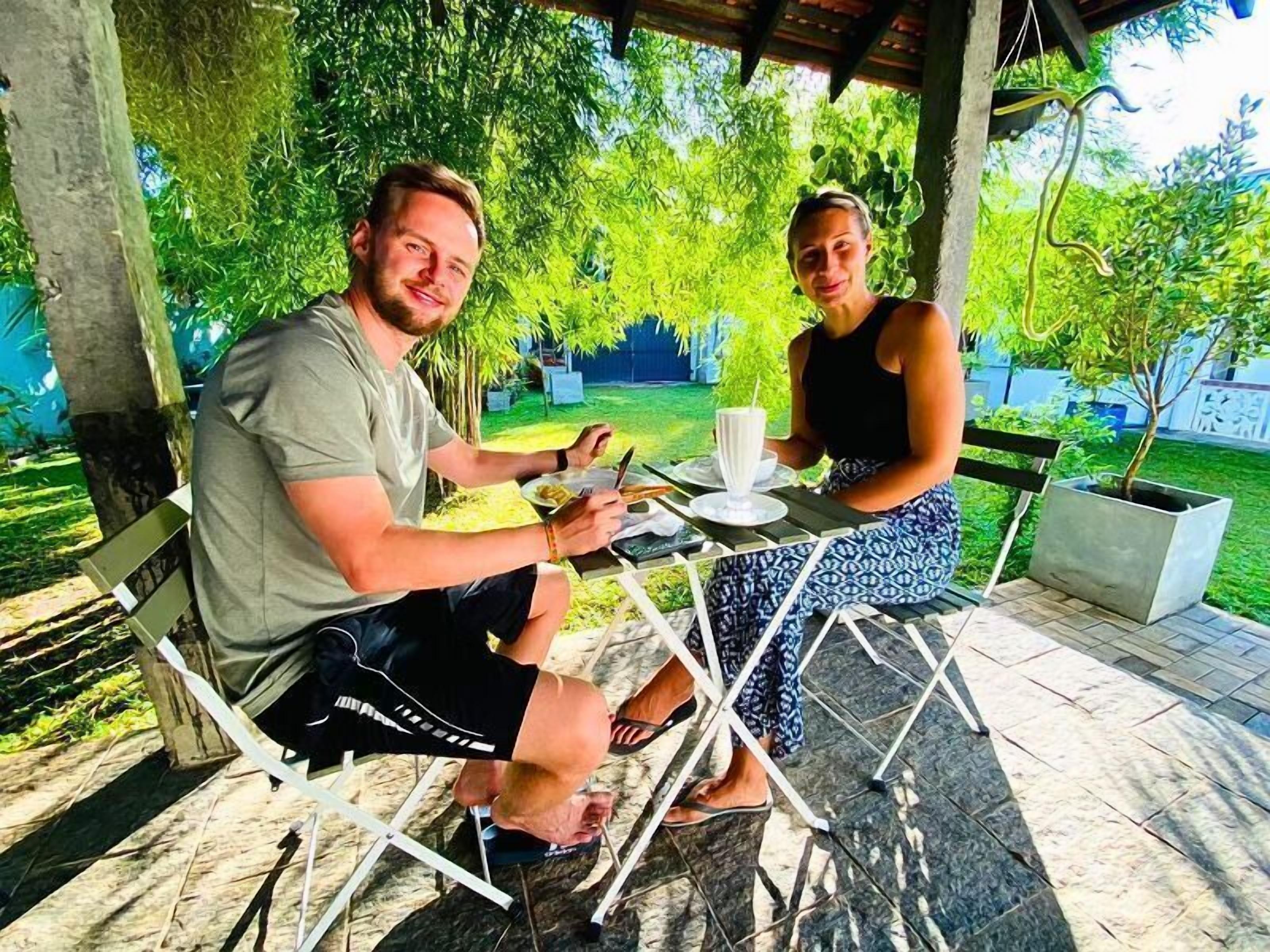 Two people seated at an outdoor table with a drink, surrounded by greenery and a covered area.
