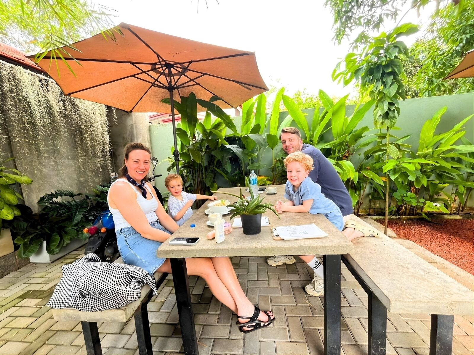 Una familia cenando en una mesa al aire libre, bajo un gran paraguas, rodeada de abundante vegetación.