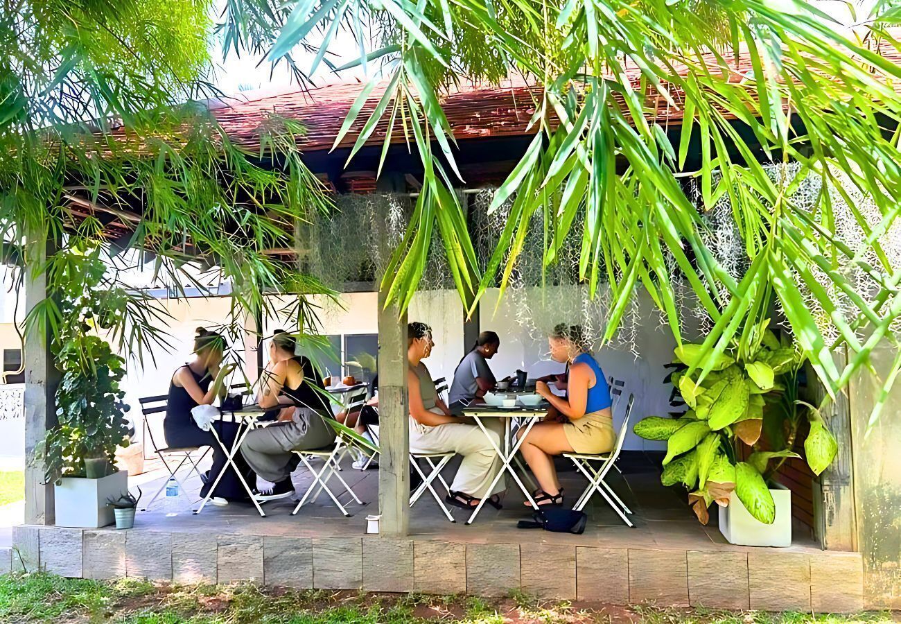 Un área de comedor al aire libre con mesas y sillas bajo una estructura cubierta, rodeada de follaje.