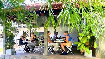 Un área de comedor al aire libre con mesas y sillas bajo una estructura cubierta, rodeada de follaje.