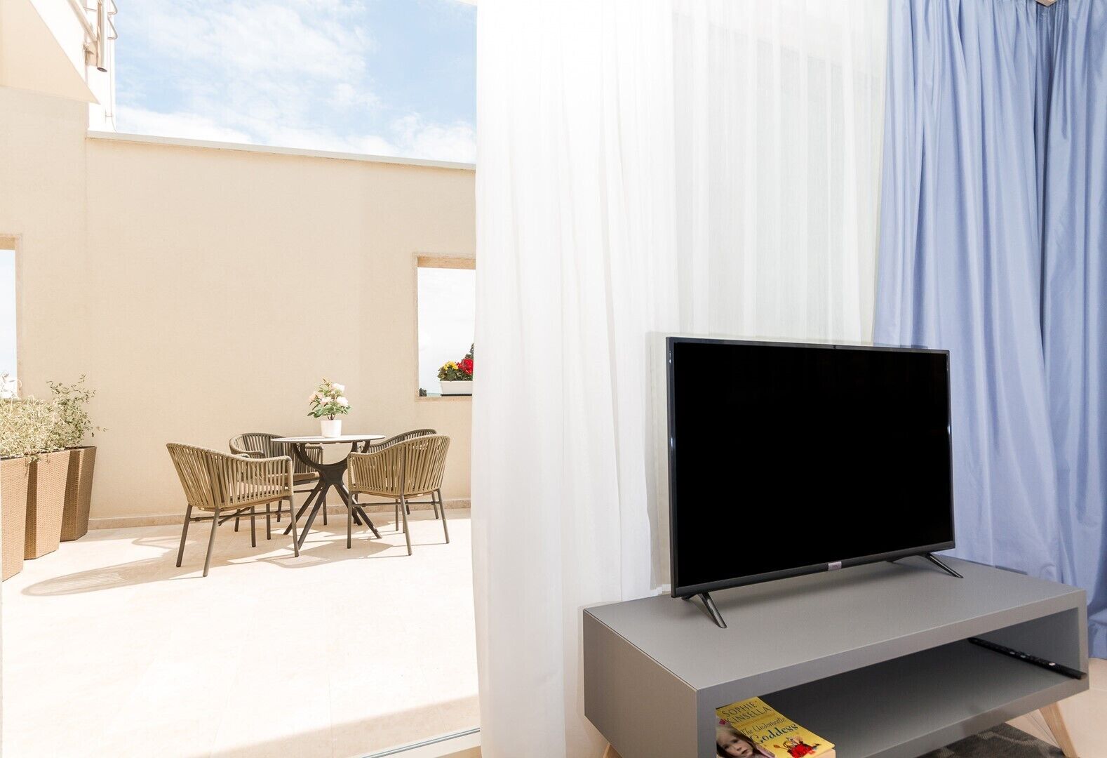 Een moderne woonkamer met een flatscreen televisie op een grijze standaard, een eethoek met rieten stoelen en een tafel, en een balkon met een wit gordijn.