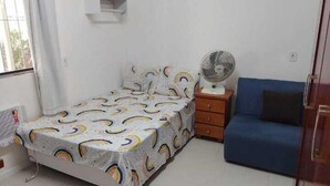 A bedroom with a bed, a bedside table with a fan, a blue sofa, and a window with curtains. - Casa das Flores (Rio das Ostras)